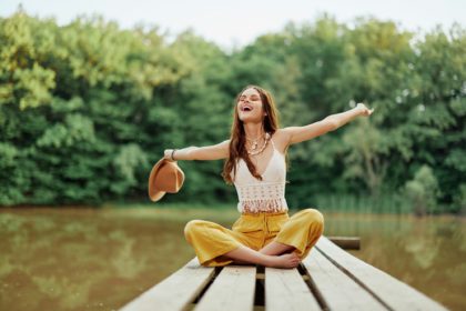 Hippie eco-activist woman traveler sits on a bridge by a lake with her arms outstretched with a hat